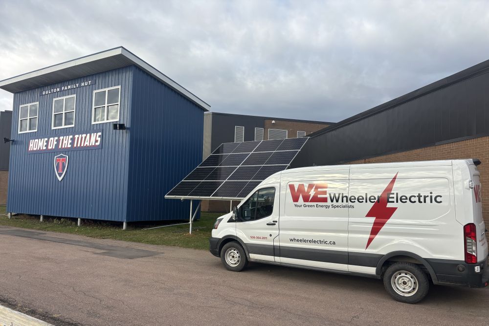 Wheeler Electric branded white Ford e-Transit van, parked next to a ground mount solar array and a blue building with the words 