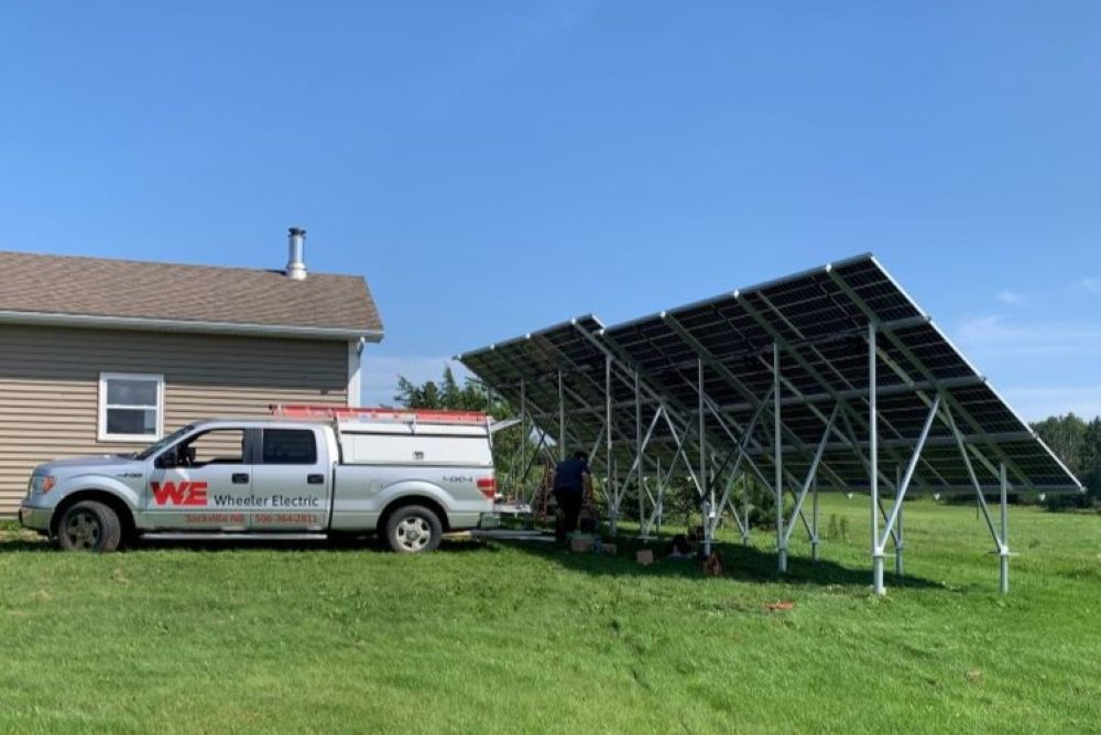 Wheeler Electric branded white work truck parked in front of a small brown bungalow and next two two ground mount solar photovoltaic arrays.  The grass is green, and the cloudless sky is blue.